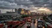 A long line of multi-axle industrial cargo trucks and containers moving slowly past a modern manufacturing unit in Chhatrapati Sambhajinagar.