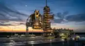 A detailed rocket stands on an illuminated launch pad at dusk against a dramatic sunset sky with a crescent moon representative image